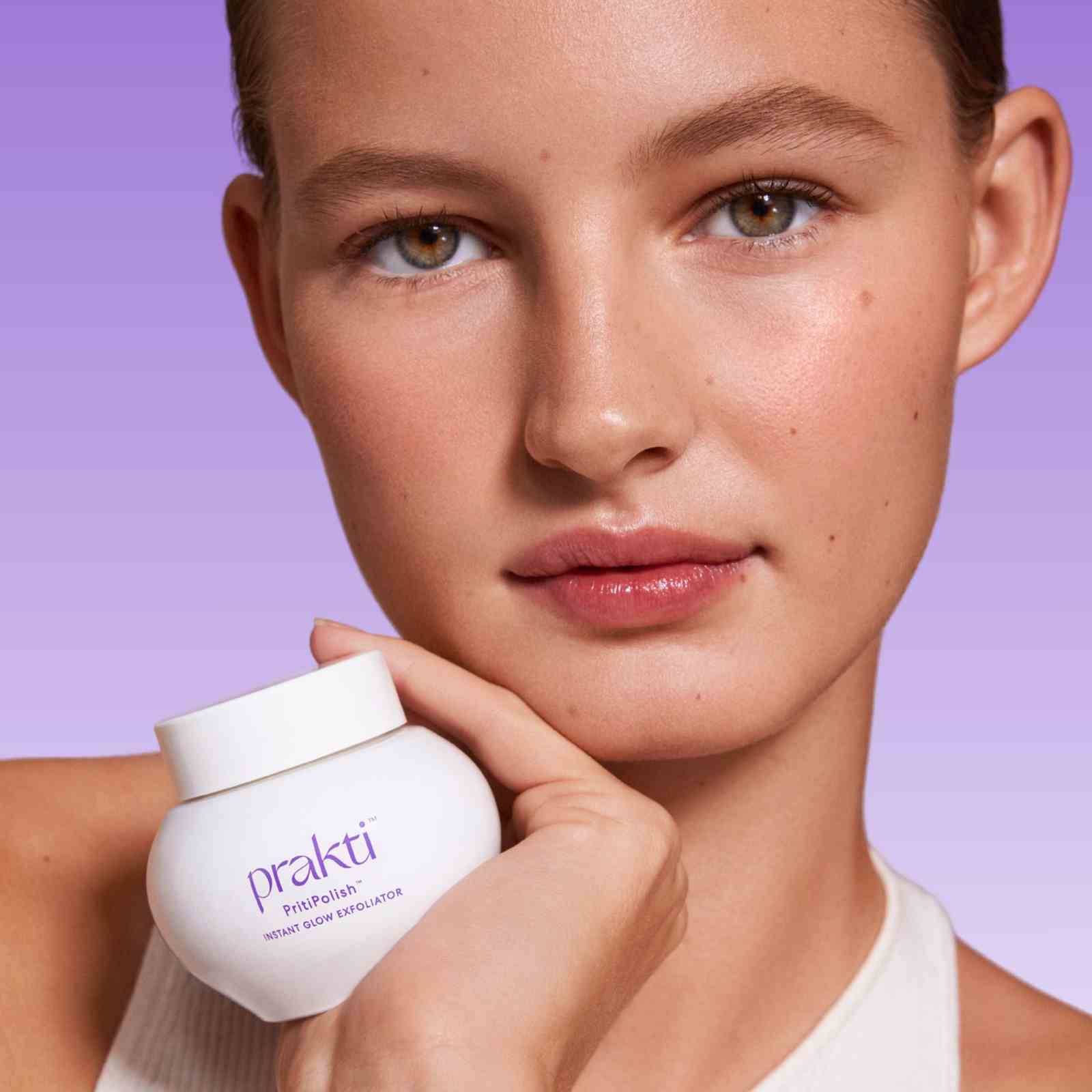woman holding PritiPolish gentle facial exfoliator jar with clear glowing skin on purple background